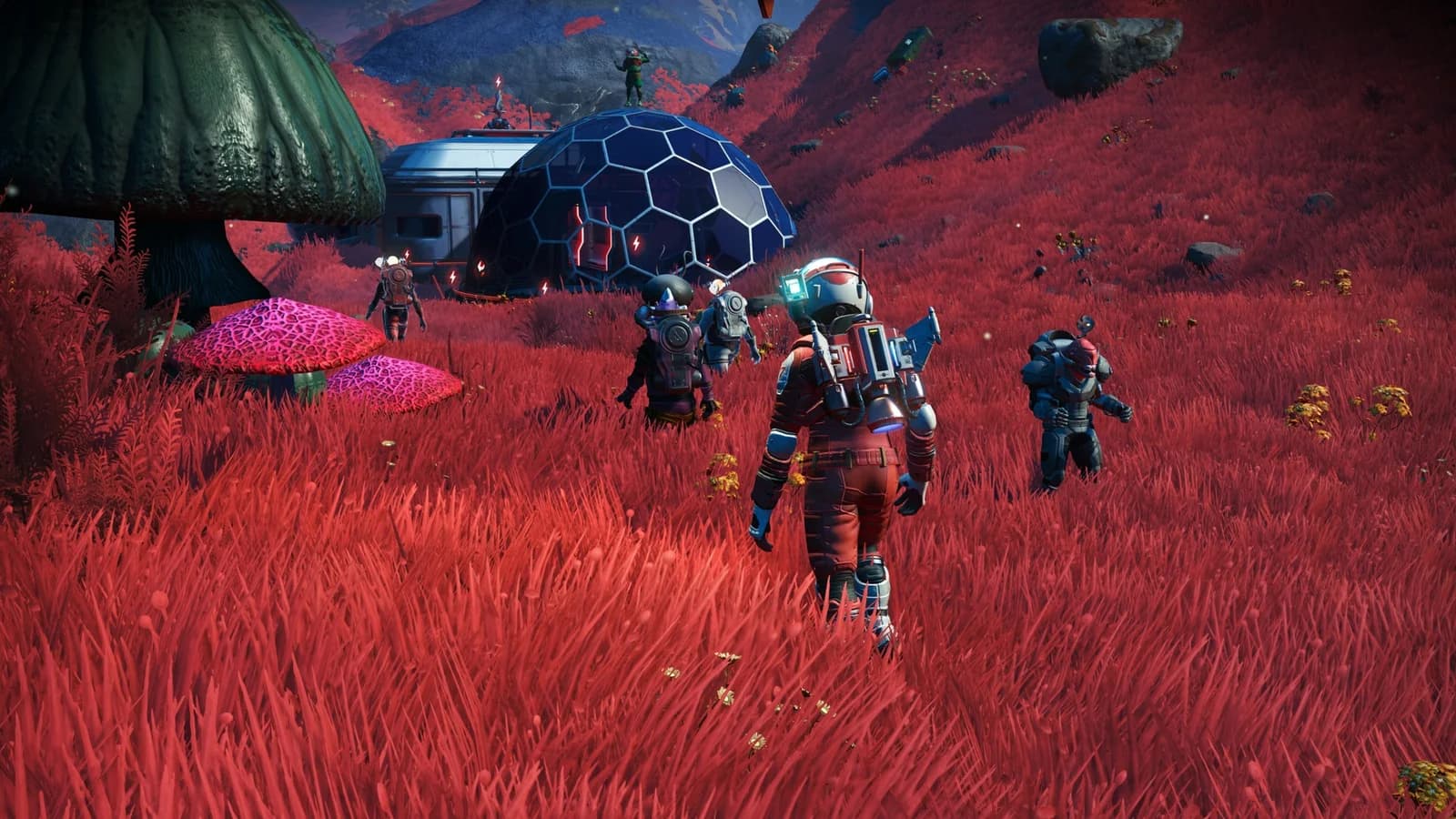 Several No Man's Sky travellers stand beside a round base module and glass dome among red grass on an alien planet.