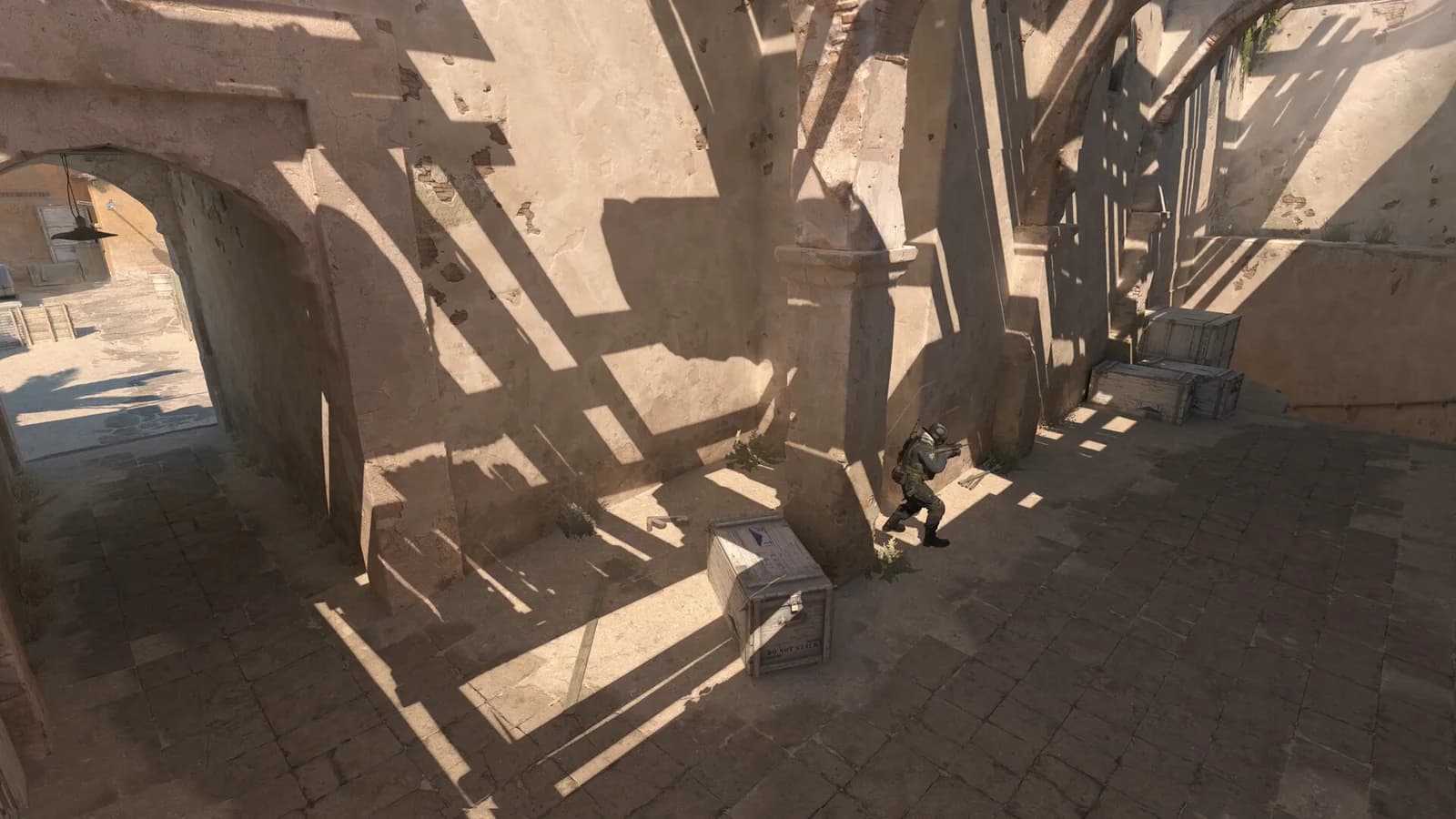 An elevated Counter-Strike 2 view of a soldier passing between tall stone walls and wooden crates.
